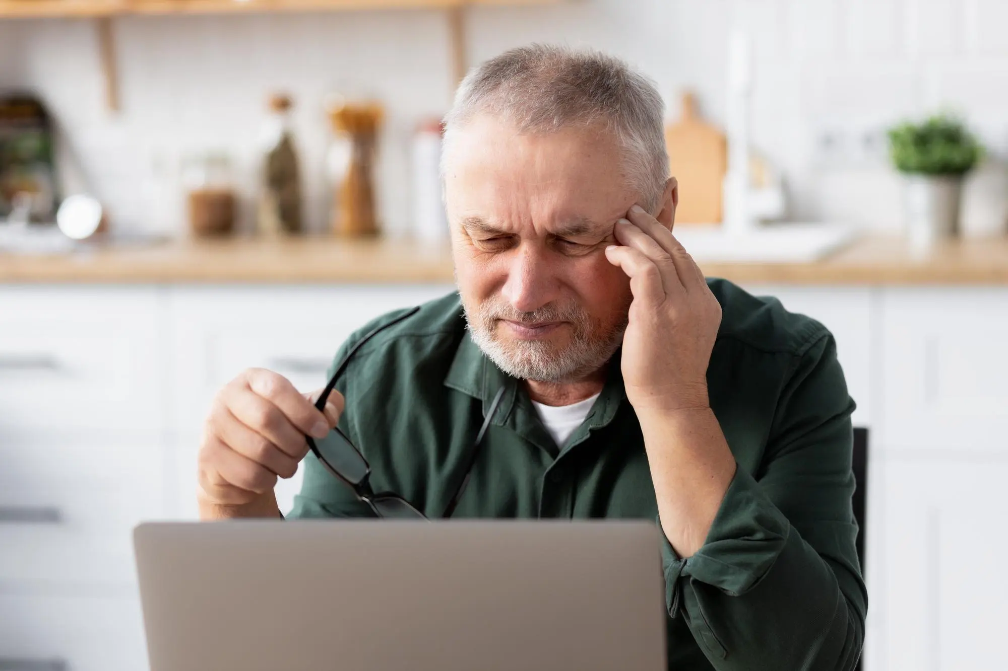 Older man experiencing a chronic headache while working on a laptop.