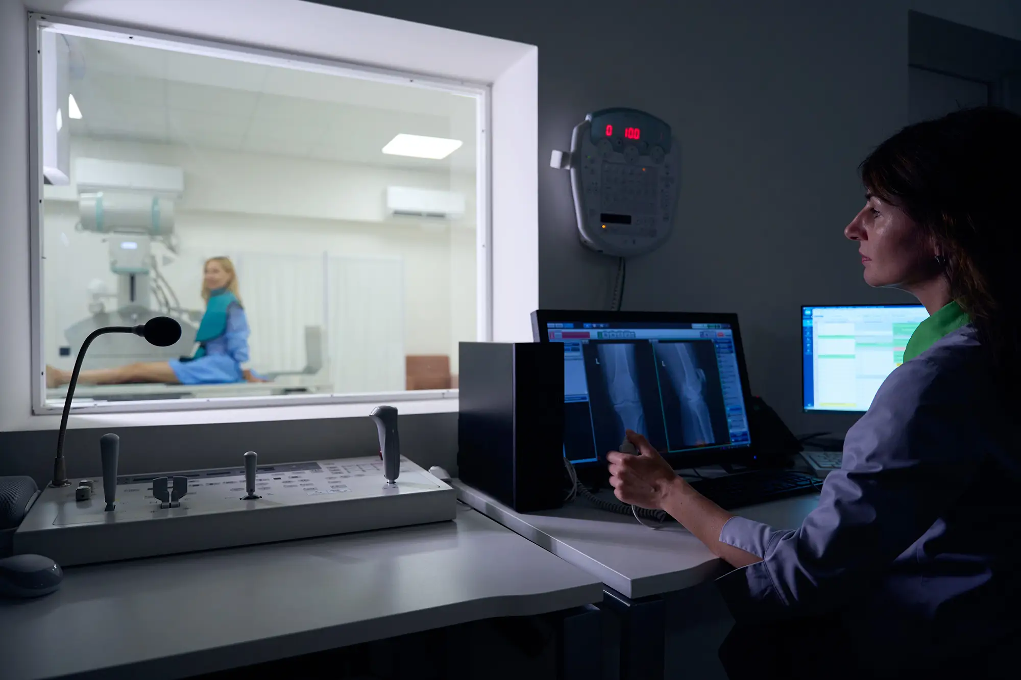 Female patient seated inside an X-ray room while a radiographer outside prepares her imaging report.