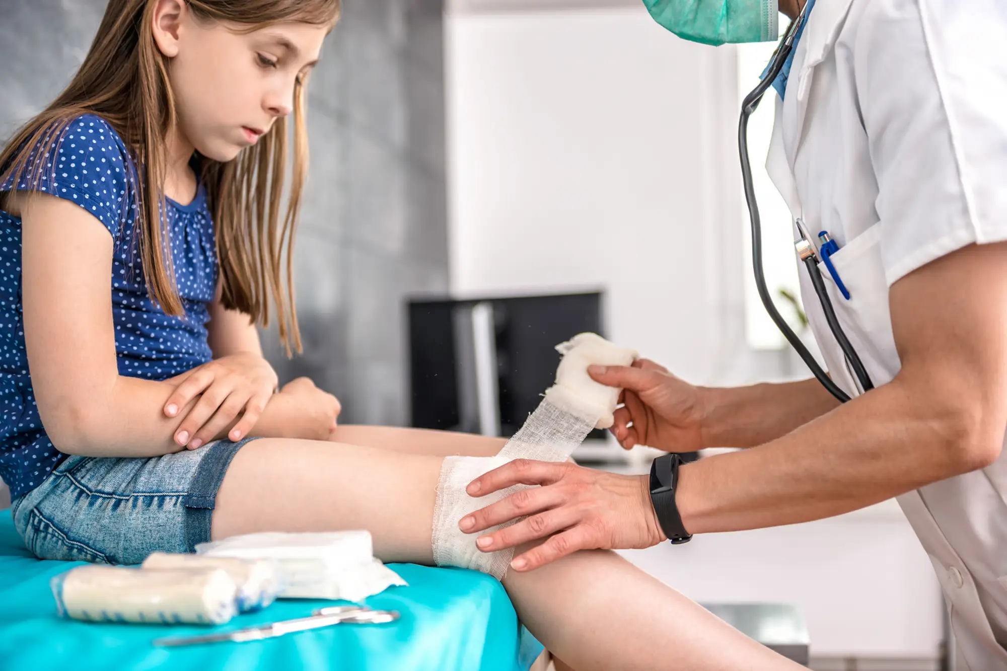 Doctor applying wound dressing to teenage girl’s injured leg joint during treatment.