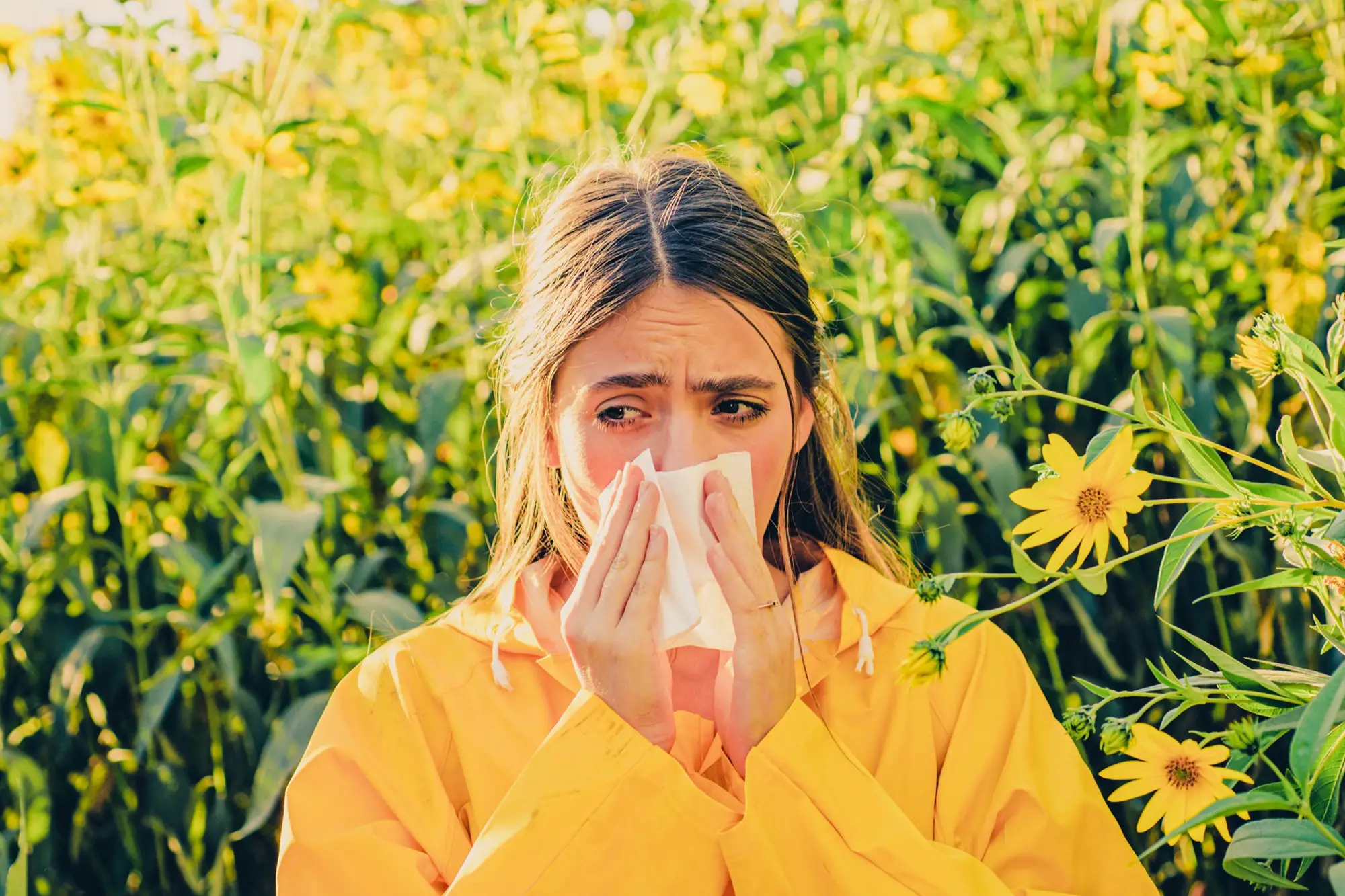 Woman covering her nose while sneezing near flowers in a green outdoor landscape.