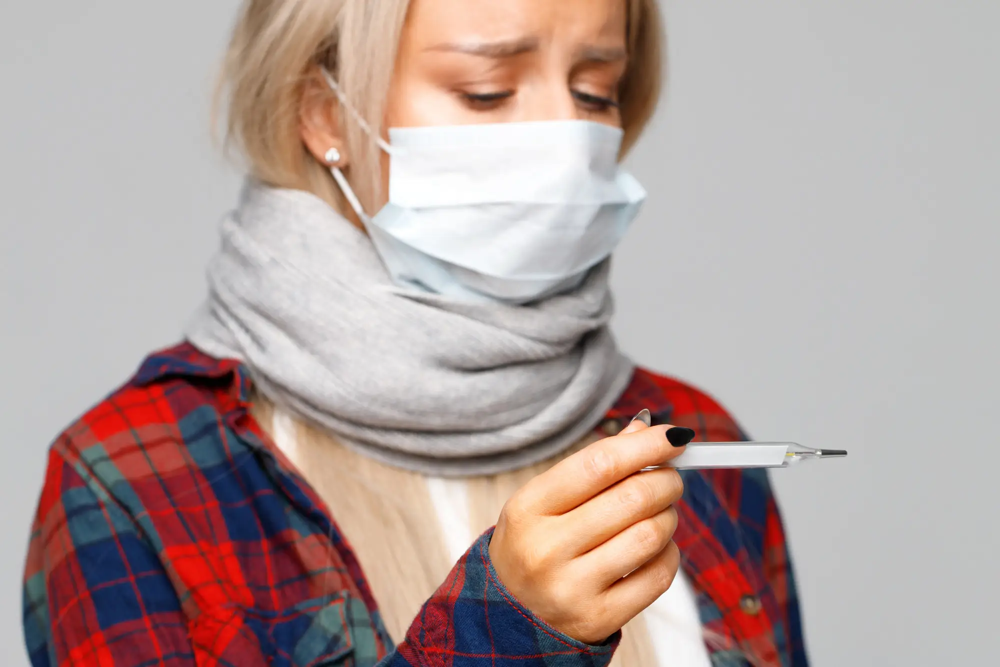Woman wearing a face mask checking her temperature with a thermometer, showing signs of flu or cold.