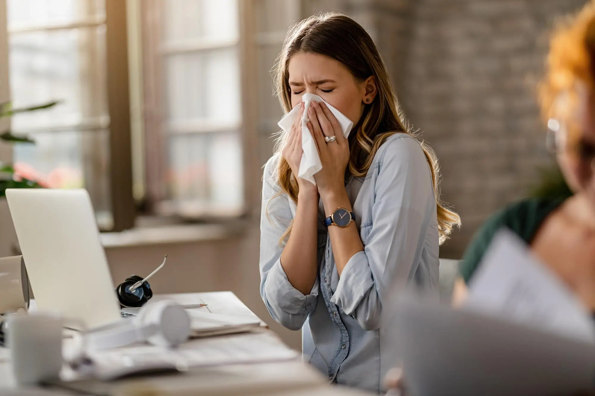 Woman using tissue paper on her nose while sneezing, showing symptoms of allergies or cold.