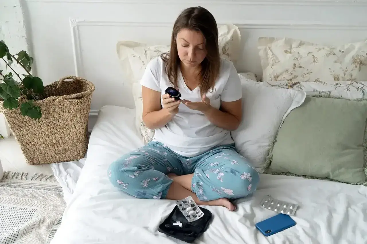 A woman sits cross-legged on a bed, wearing a white t-shirt and light blue patterned pajama pants, checking her blood sugar with a glucometer.