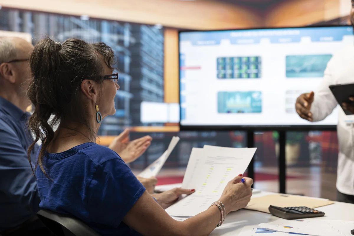 Older adults holding printed charts and watching a presenter point to data on a large screen