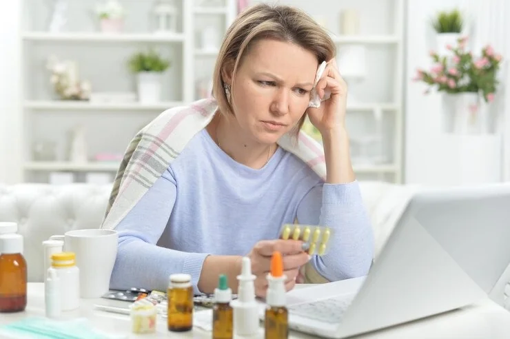 Woman at a laptop holding a blister pack of medication with a tissue at her temple, showing fatigue from hormone imbalance