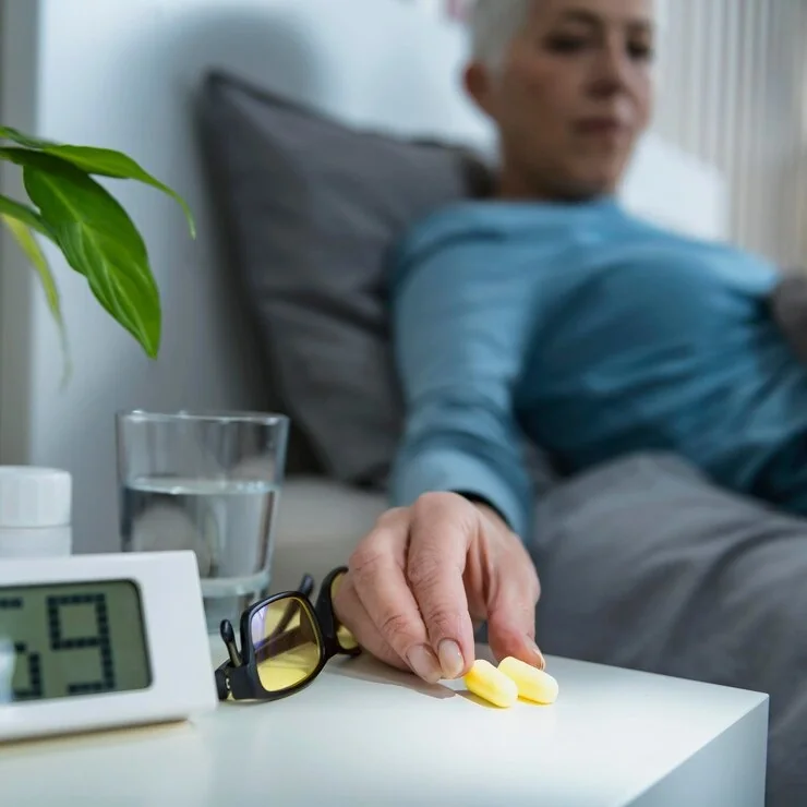 Mature woman resting in bed reaching for earplugs on a nightstand beside a glass of water and a digital clock