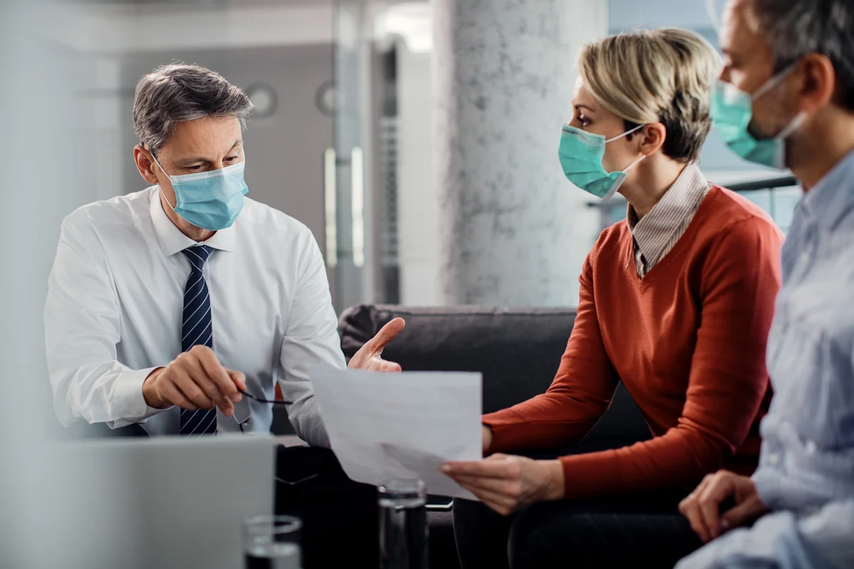 Man in a white shirt and tie explaining paperwork to a woman and man, all wearing face masks, during a private consultation