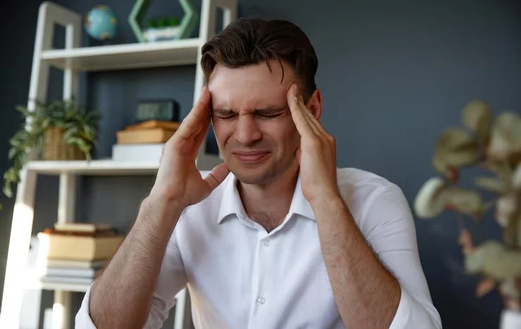 Man at a desk holding both hands to his temples, showing a tension headache caused by stress