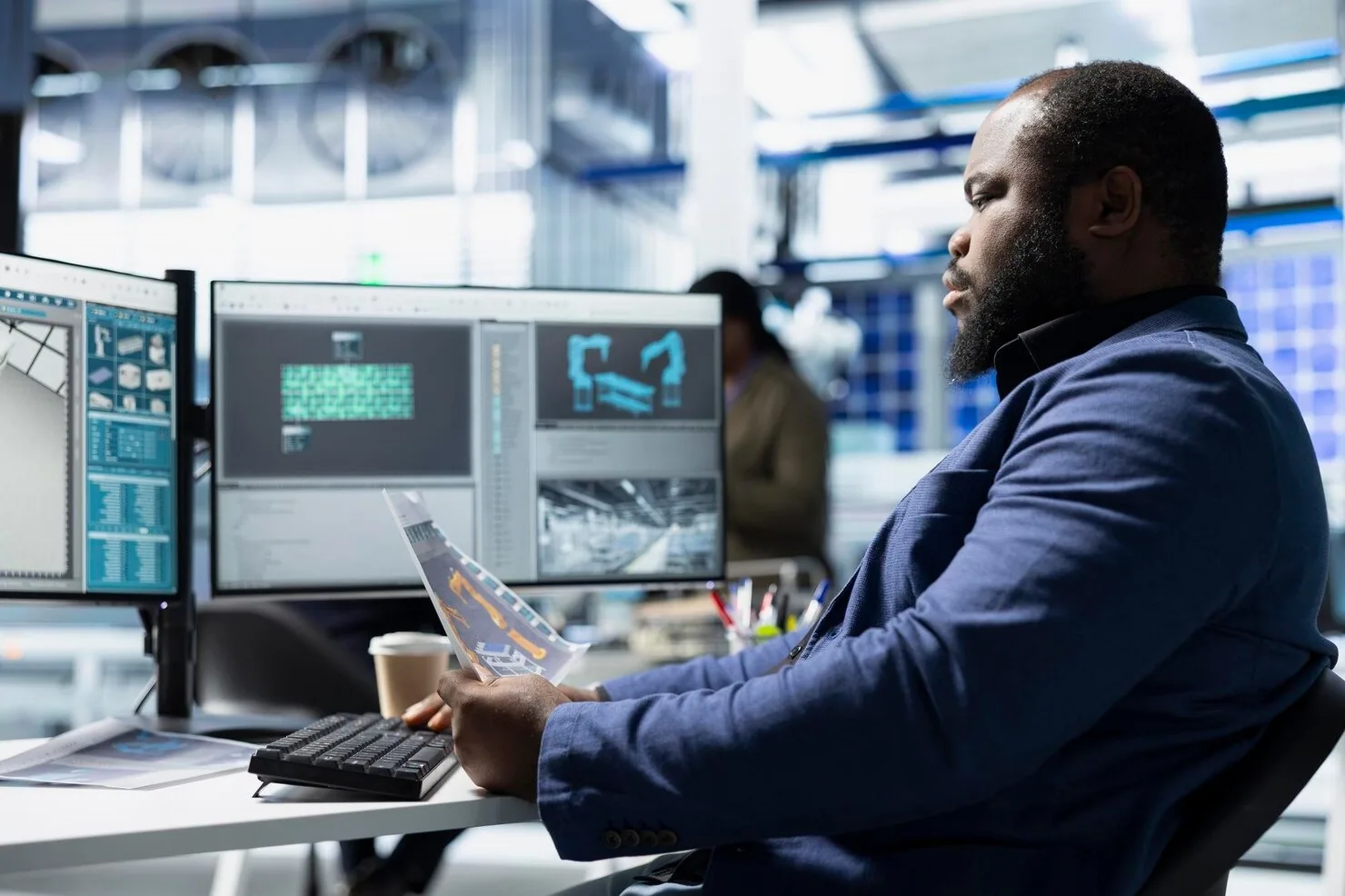 Professional in a navy blazer reviewing data on dual monitors and a printed report at a desk in a modern workspace
